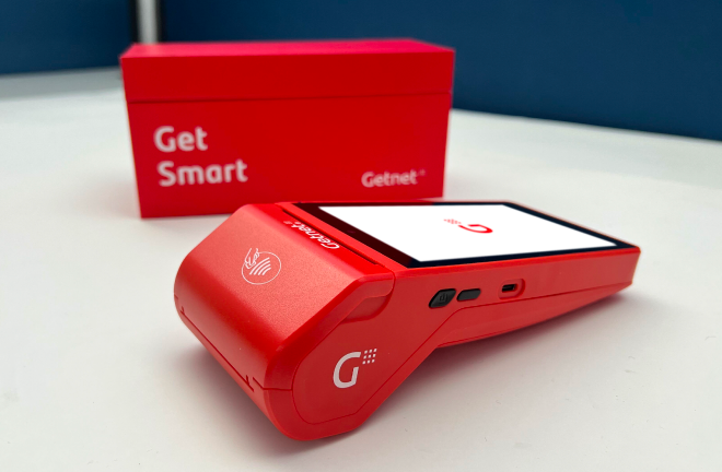 Terminal de cobro Getnet roja apoyada sobre superficie blanca, con pantalla encendida mostrando el logo y caja roja detrás con texto 'Get Smart'.