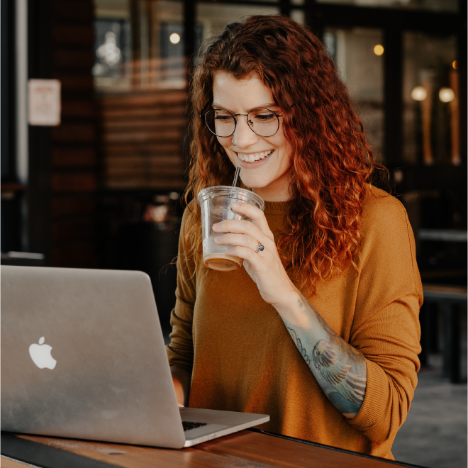Persona sonriendo mientras trabaja en una laptop y sostiene un vaso transparente con bebida, sentada en una mesa de madera en un ambiente interior.