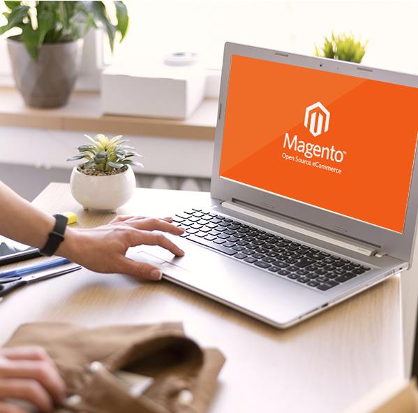 Mano usando una laptop con pantalla que muestra el logo de Magento y la frase “Open Source eCommerce”, mientras la otra mano está apoyada sobre una prenda doblada, sobre un escritorio con una planta y objetos de oficina.
