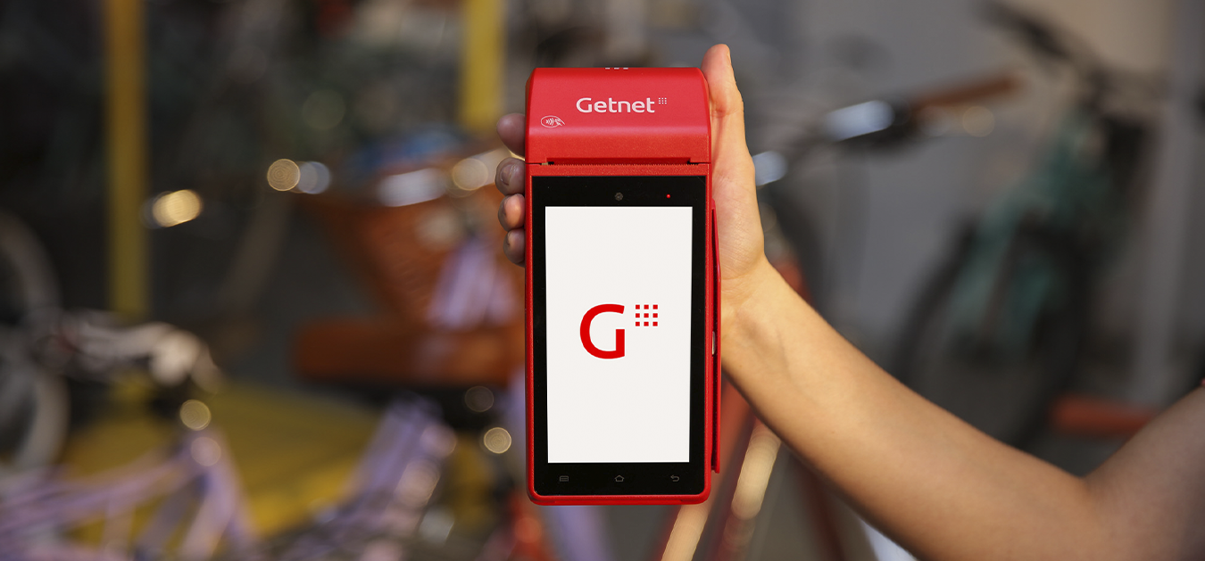 Mano sosteniendo una terminal de cobro Getnet roja con pantalla encendida mostrando el logo, sobre fondo difuminado.