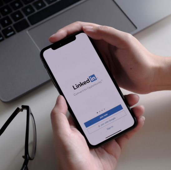 Teléfono móvil mostrando la pantalla de inicio de sesión de LinkedIn, sostenido con ambas manos sobre un escritorio con una computadora portátil y gafas.