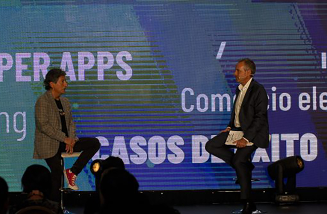 Dos personas sentadas en un escenario durante un evento, con fondo que muestra palabras como 'Súper Apps', 'Casos de éxito' y 'Comercio electrónico'.