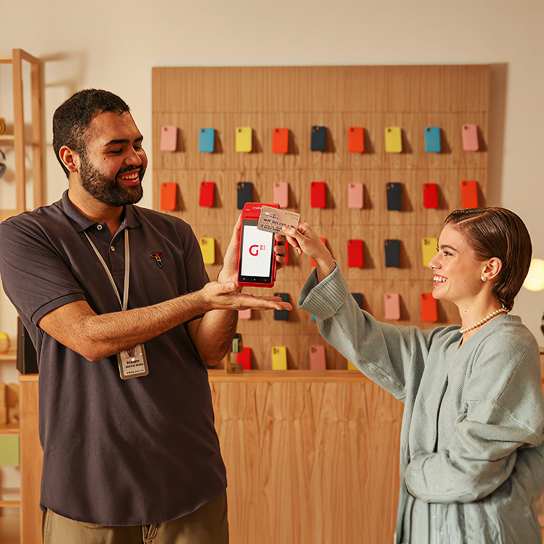 Dos personas en una tienda: una sostiene una terminal GetPOS con el logo de Getnet en la pantalla y la otra apoya una tarjeta en la terminal. Al fondo se ven fundas de celular colgadas en la pared.