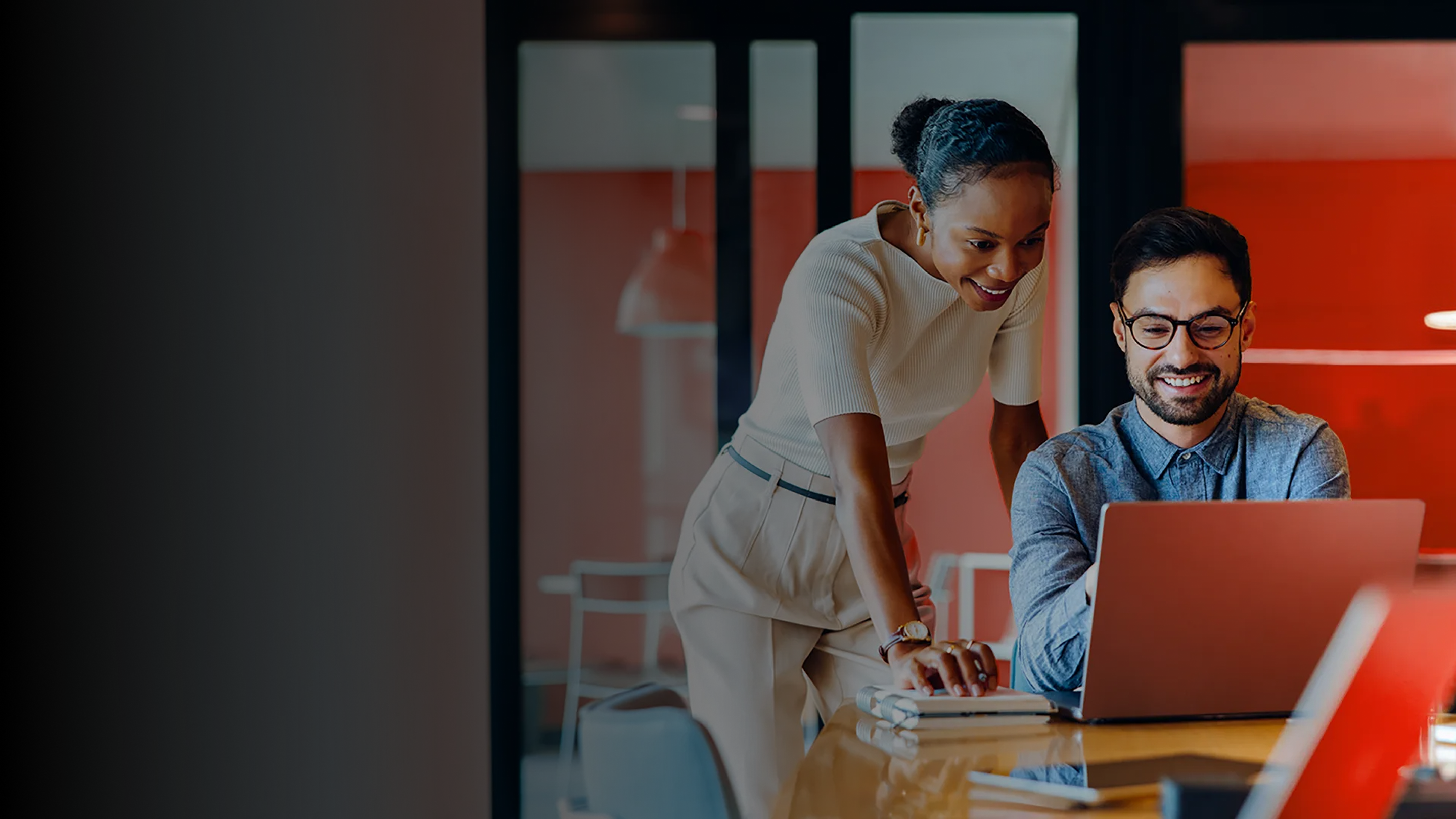 Dos personas en una oficina; una está de pie junto a un escritorio y otra trabaja en una computadora portátil.