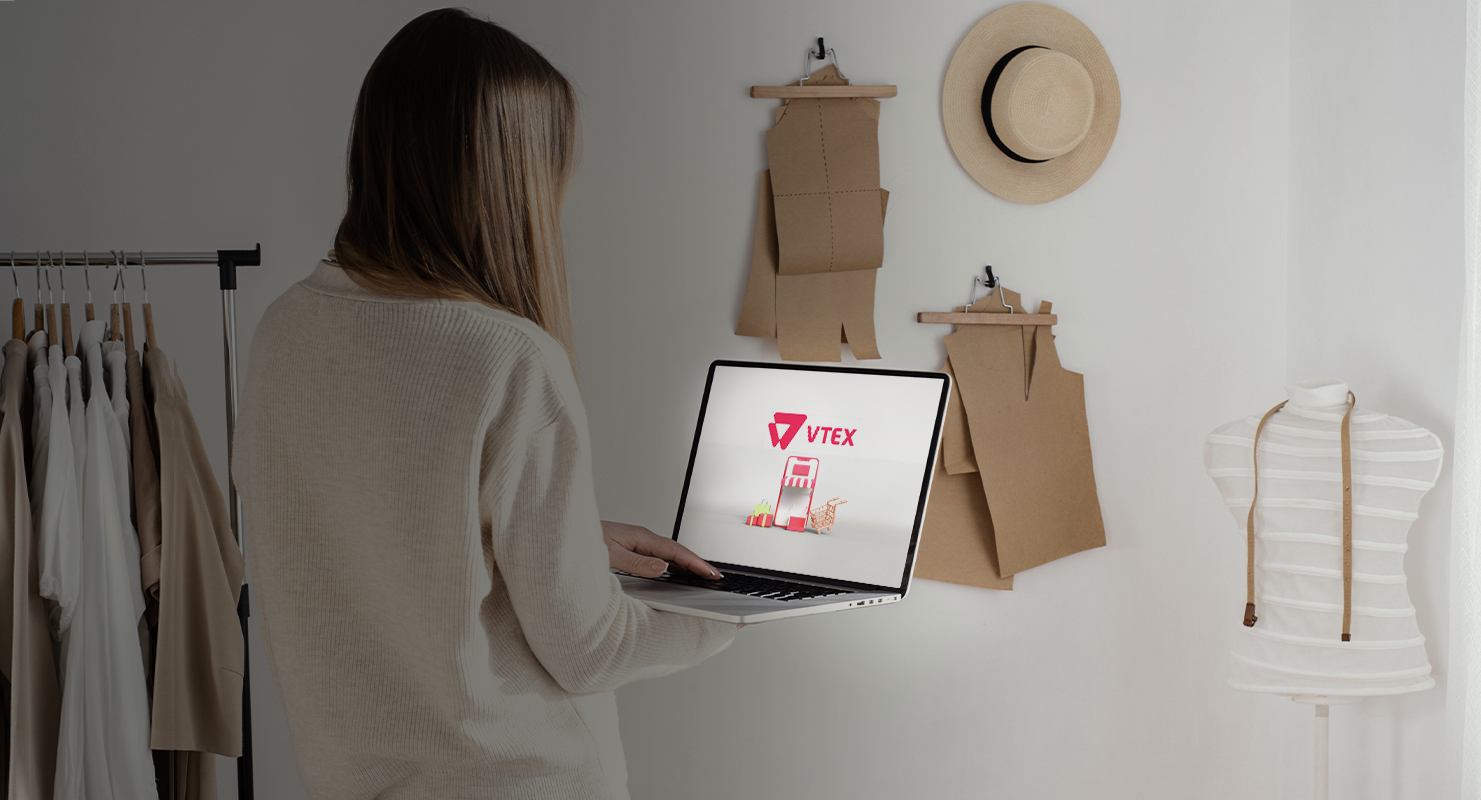 Persona sosteniendo una laptop que muestra el logo de VTEX, en una habitación con prendas colgadas, sombrero en la pared y un maniquí.